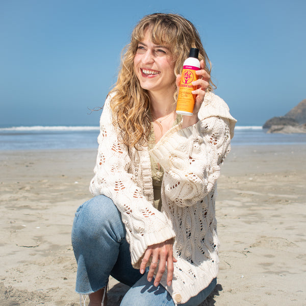 Woman on a beach holding a bottle of Jessicurl Confident Coils Styling Solution